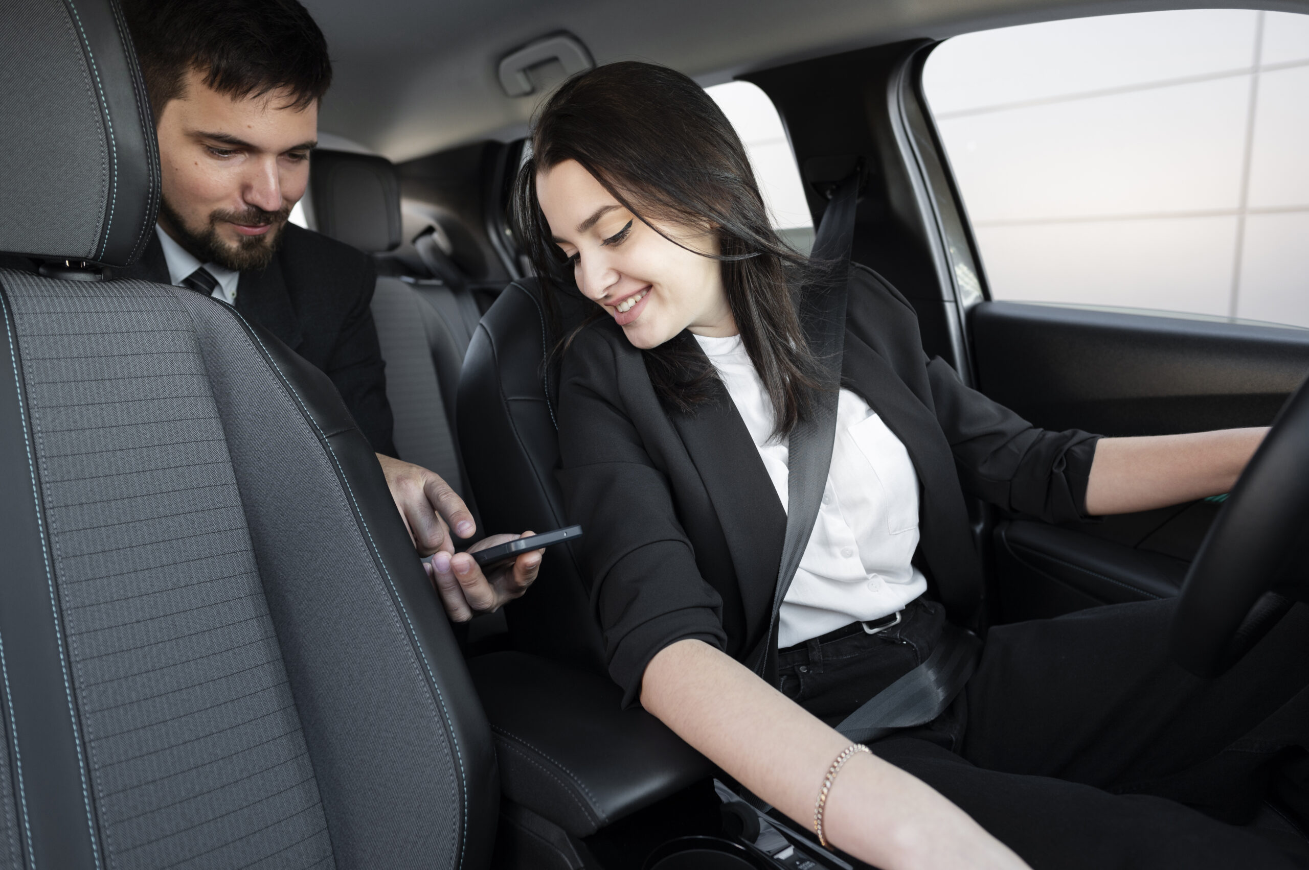 Modern bir otomobilin içinde, sürücü koltuğunda oturan emniyet kemeri bağlı bir kadın ve arka koltuktaki erkek yolcunun birlikte bir akıllı telefon ekranına bakarak gülümsedikleri anın iç çekimi.